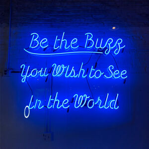 A blue neon sign reading be the buzz you wish to see in the world