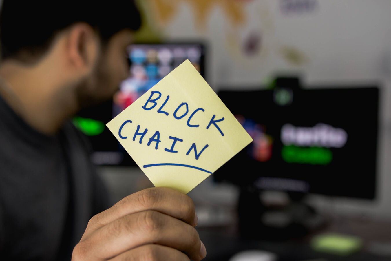 An IT developer holding up a yellow sticky note with blockchain written on it in blue ink