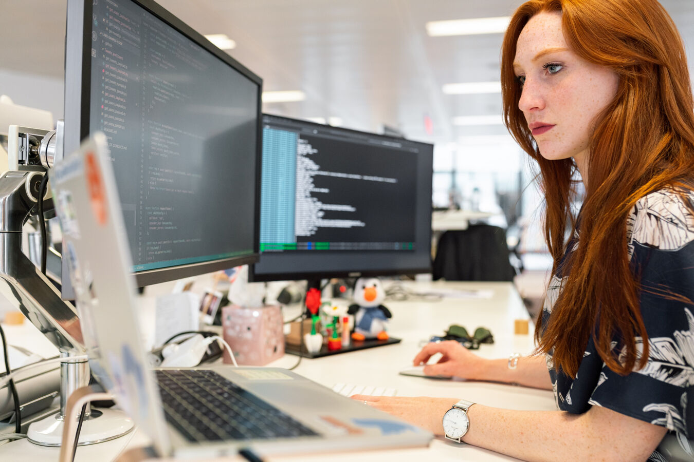 A young woman sitting coding at multiple screens in an office setting