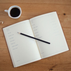 A cup of black coffee, diary and pencil sitting on a wooden desktop