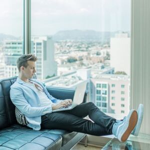 kannettavaa tietokonetta käyttävä nuori mies kattohuoneistossa, josta on näköala kaupunkiin