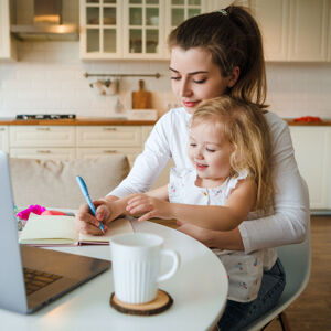 Businesswoman wrting in a notbook, with a child on her knee sitting at the kitchen table with a computer and mug