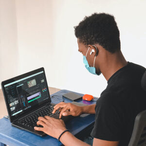 Young black man wearing a blue facemask working at a laptop