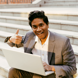 Young Indian man in a white shirt and a business suit sitting on some steps with a laptop, smiling and typing