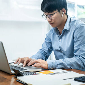 A businessman is typing on a laptop while in a virtual meeting. Notepads and a mobile phone are in the foreground