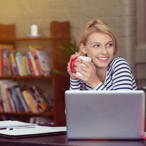 Smiling young woman taking a coffee break as she sits at her laptop working from her home office
