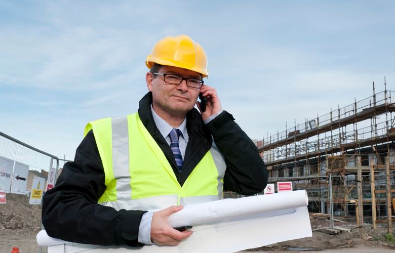 A project manager standing on a building site wearing a yellow hard hat and reflective vest while on a mobile phone